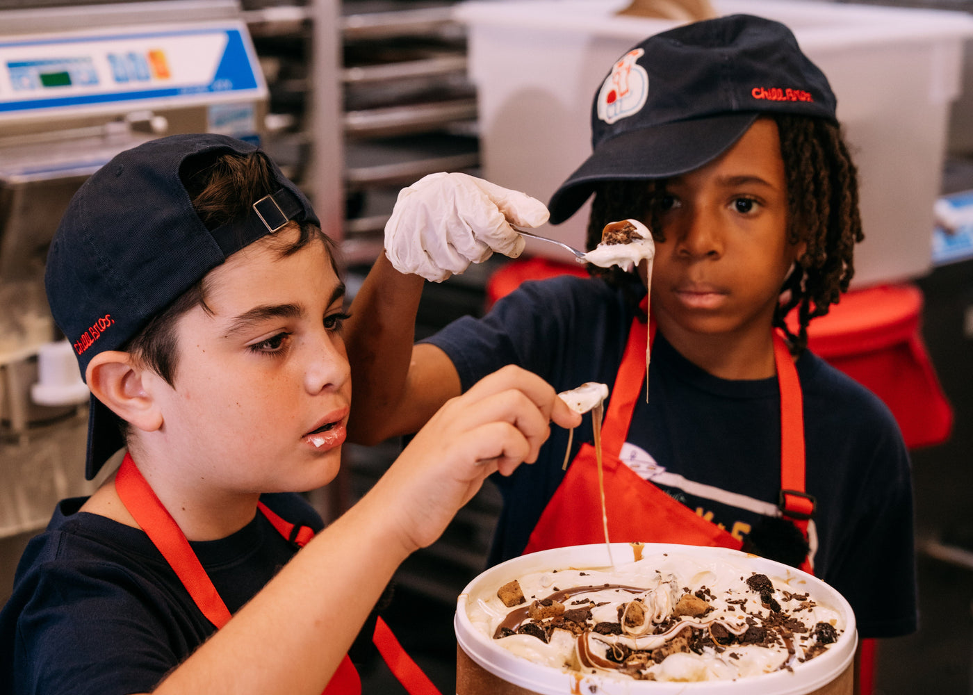 Two children wearing matching red aprons and black caps with a logo are serving ice cream from a large brown paper cup in a commercial kitchen setting.