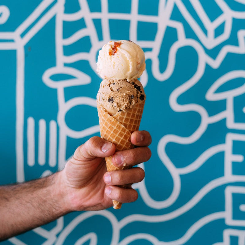 A hand holding a double-scoop ice cream cone with vanilla and chocolate chip ice cream in front of a bright blue graffiti-patterned background.