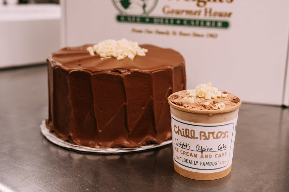 A chocolate cake and a branded cup of Wright's Gourmet House alpine Cake & ice Cream on a stainless steel counter, with the brand's sign visible in the background.